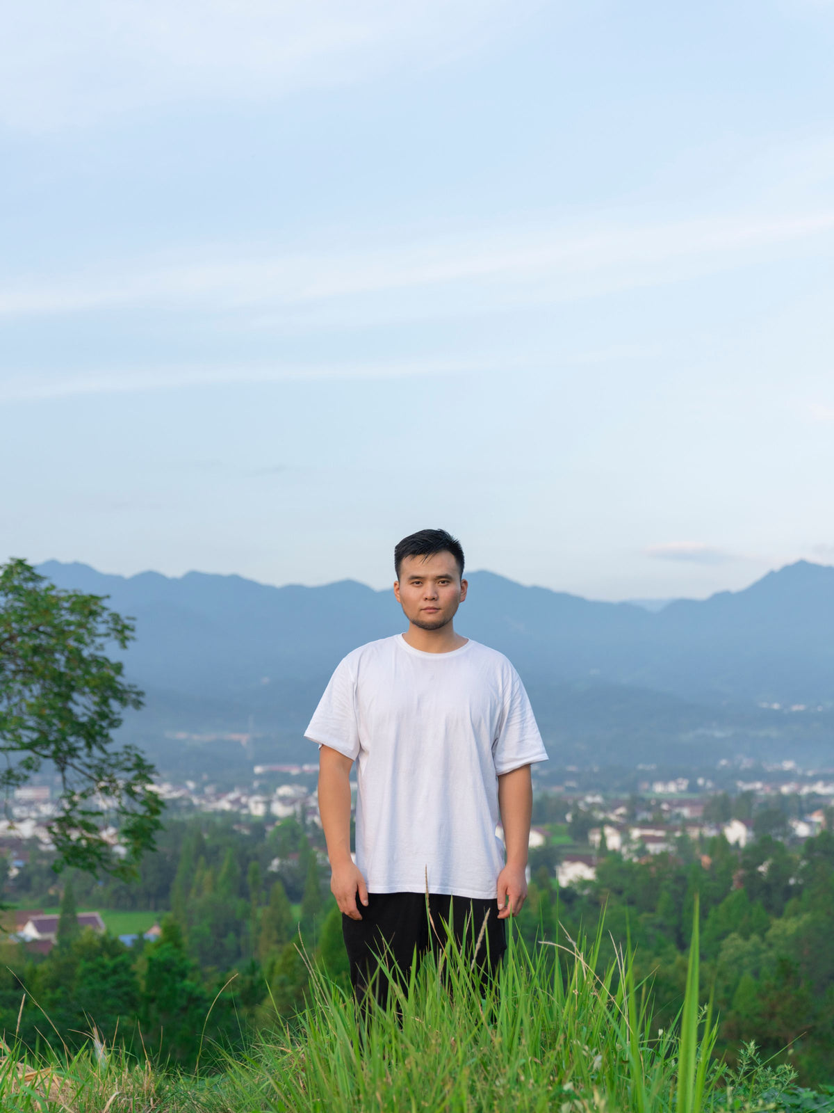Man in a plain white T-shirt outdoors with green hills behind him