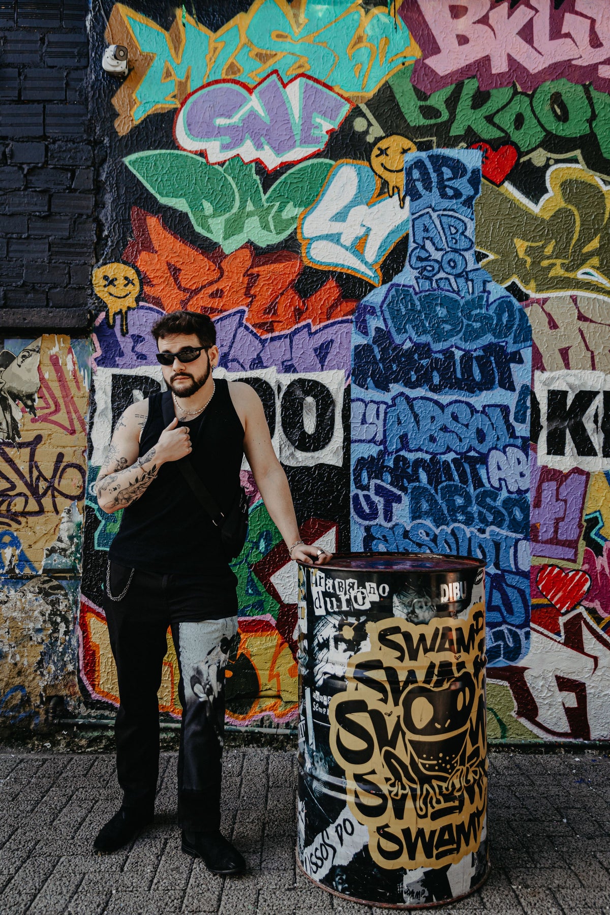 Tattooed man in black streetwear beside a colourful graffiti wall